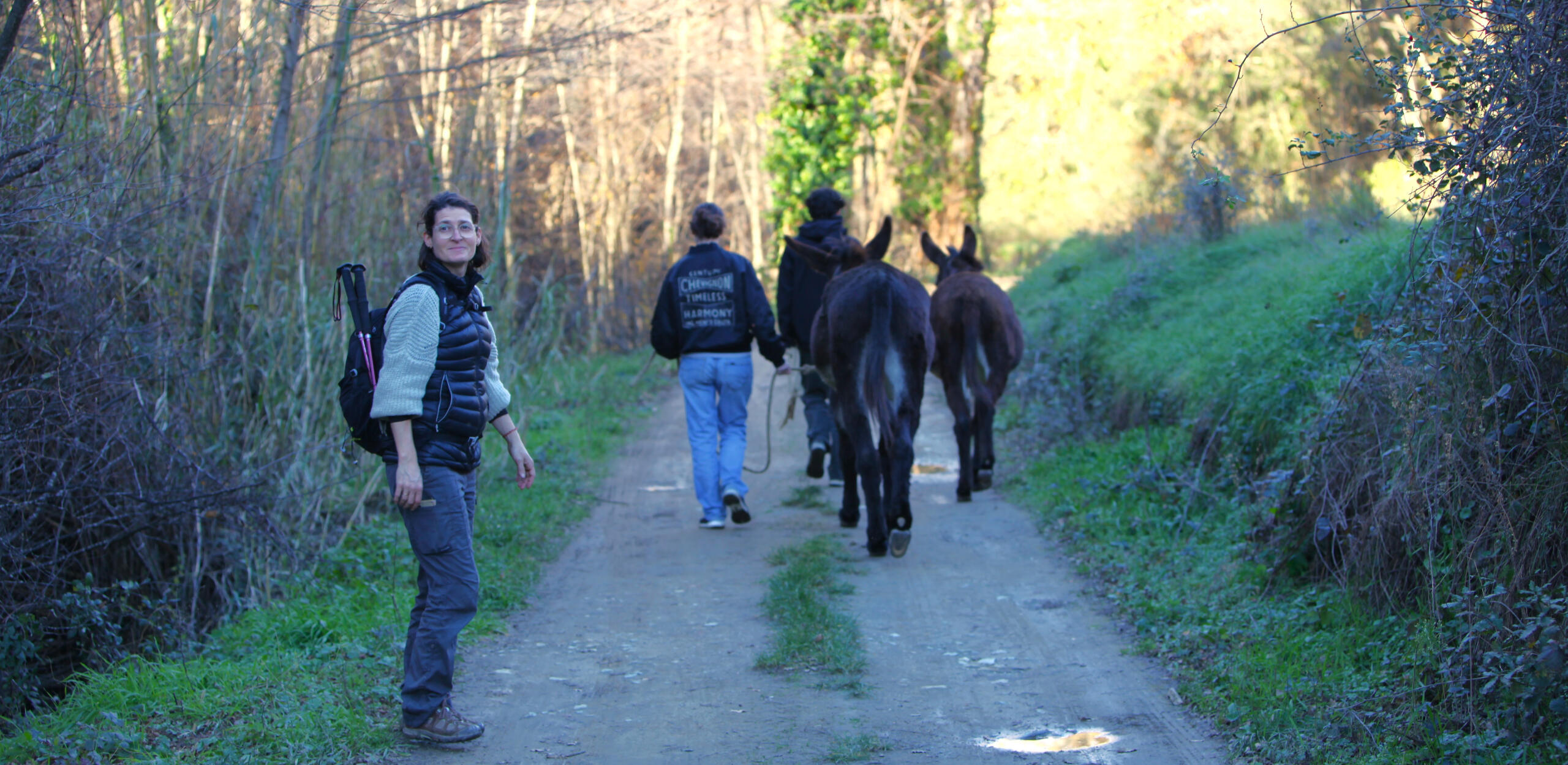 Emmanuelle en accompagnement d'ados par la médiation asine. Sur un chemin de campagne, des adolescents accompagnés d'ânes marchent. L'intervenante en médiation animale, se retourne souriante.